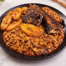 A bowl of Nigerian brown beans porridge with fish, beside a few slices of boiled plantain.