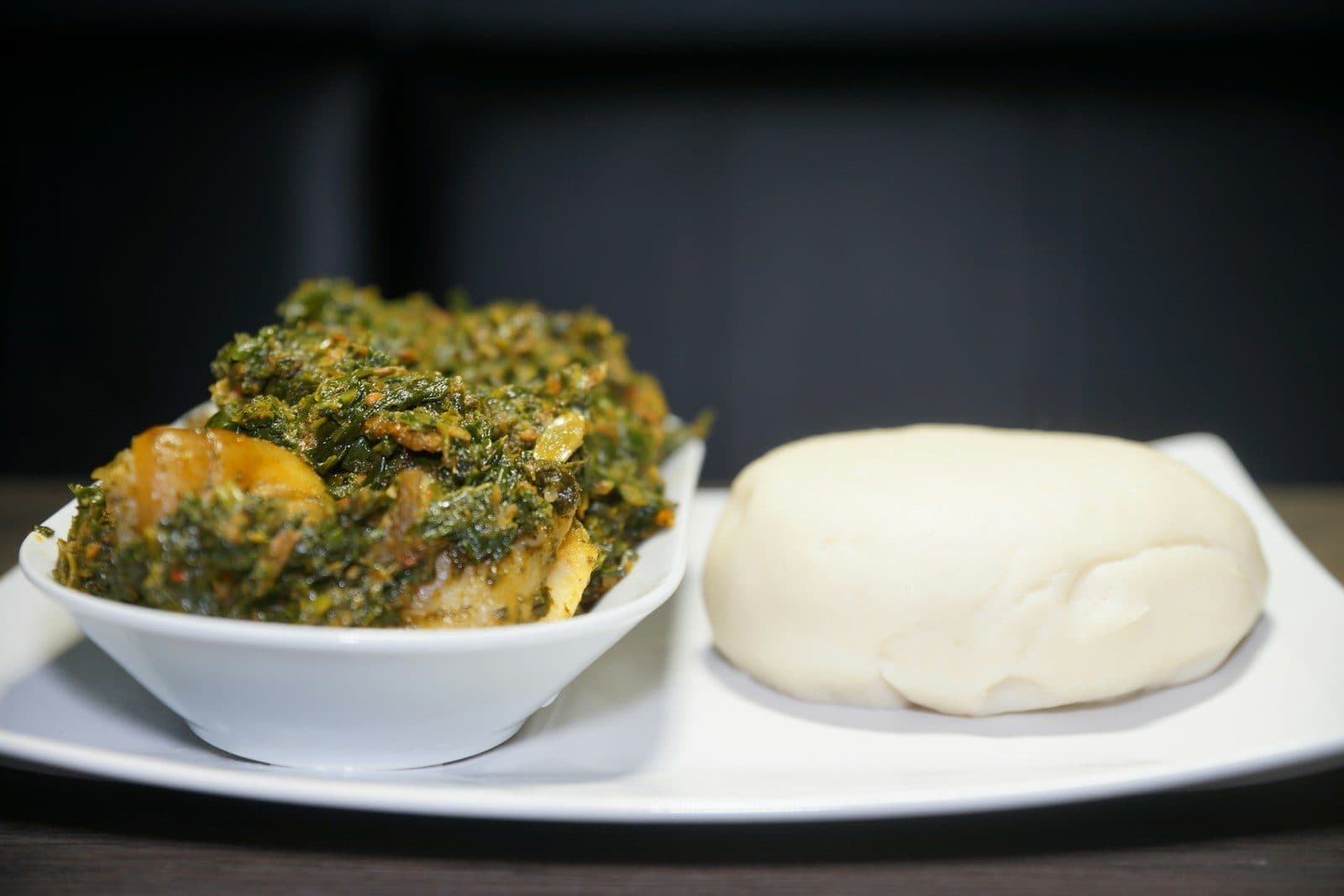A bowl of green leafy vegetable soup beside a ball of white swallow on a white plate